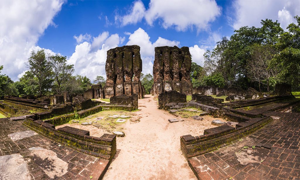 The ruins of a grand ancient palace complex in Polonnaruwa, Sri Lanka. The central feature is a pair of towering, partially collapsed structures that once formed the entrance to the palace. The surrounding area is a mix of overgrown trees, stone pathways, and remnants of other buildings.