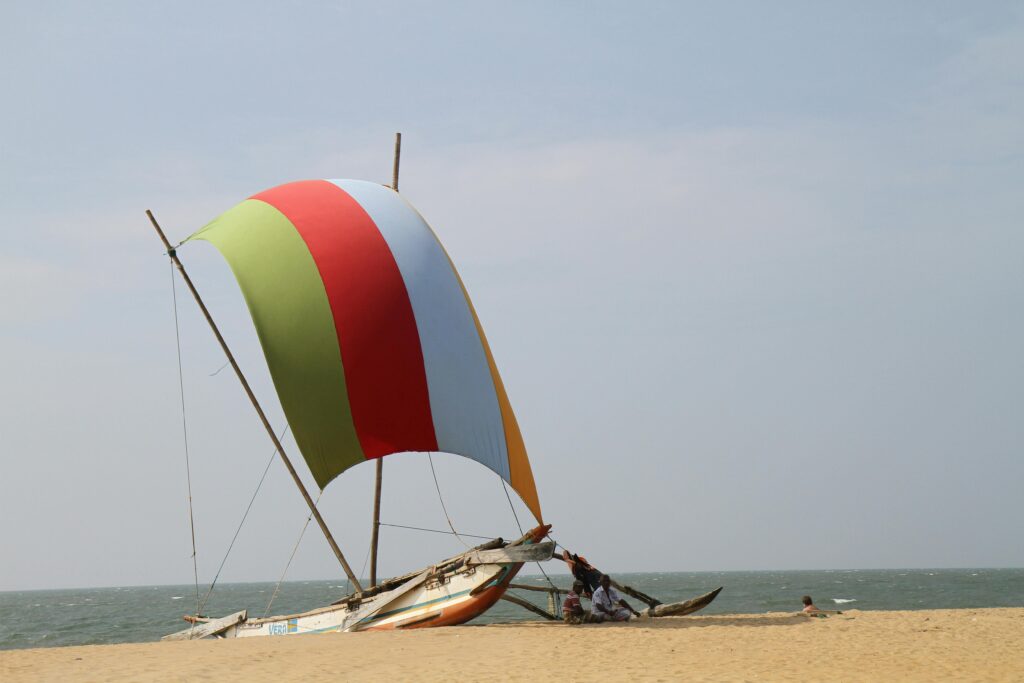 A colorful sailboat with green, red, and white stripes on Bentota Beach, Sri Lanka. The boat, a traditional Sri Lankan outrigger canoe known as an Oruwa, is set against a backdrop of golden sand and a calm blue sea under a clear sky. Another smaller boat and a person sitting nearby suggest leisure activities or local fishing practices. This image showcases the serene beauty of Bentota Beach, perfect for a relaxing beach vacation arranged by Deluxe Holidays.