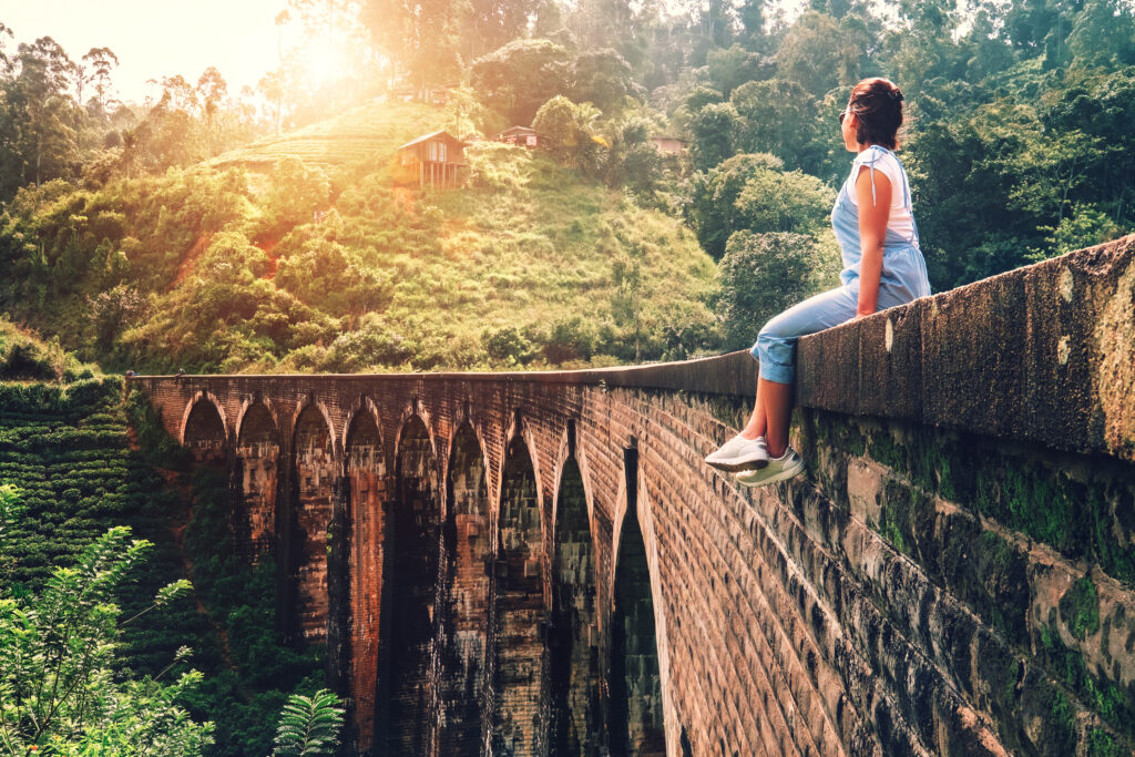 Plan your holidays to Sri Lanka for a limited time with different activities, and adventures with Delux Holidays, a reliable tour operators to Sri Lanka. A scenic view in Ella, Sri Lanka, possibly related to a railway bridge. The famous Nine Arches Bridge, also known as the Demodara Bridge, is in Ella. A person is sitting on the edge of this historic stone bridge, which features nine elegant arches. The bridge is surrounded by lush greenery, with dense foliage and trees in the background.