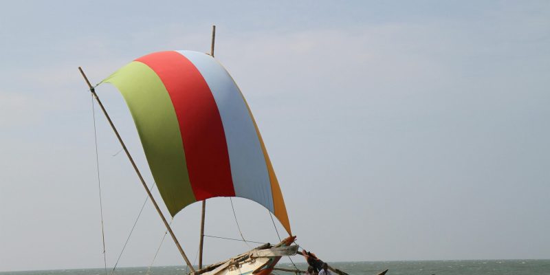 A colorful sailboat with green, red, and white stripes on Bentota Beach, Sri Lanka. The boat, a traditional Sri Lankan outrigger canoe known as an Oruwa, is set against a backdrop of golden sand and a calm blue sea under a clear sky. Another smaller boat and a person sitting nearby suggest leisure activities or local fishing practices. This image showcases the serene beauty of Bentota Beach, perfect for a relaxing beach vacation arranged by Deluxe Holidays.